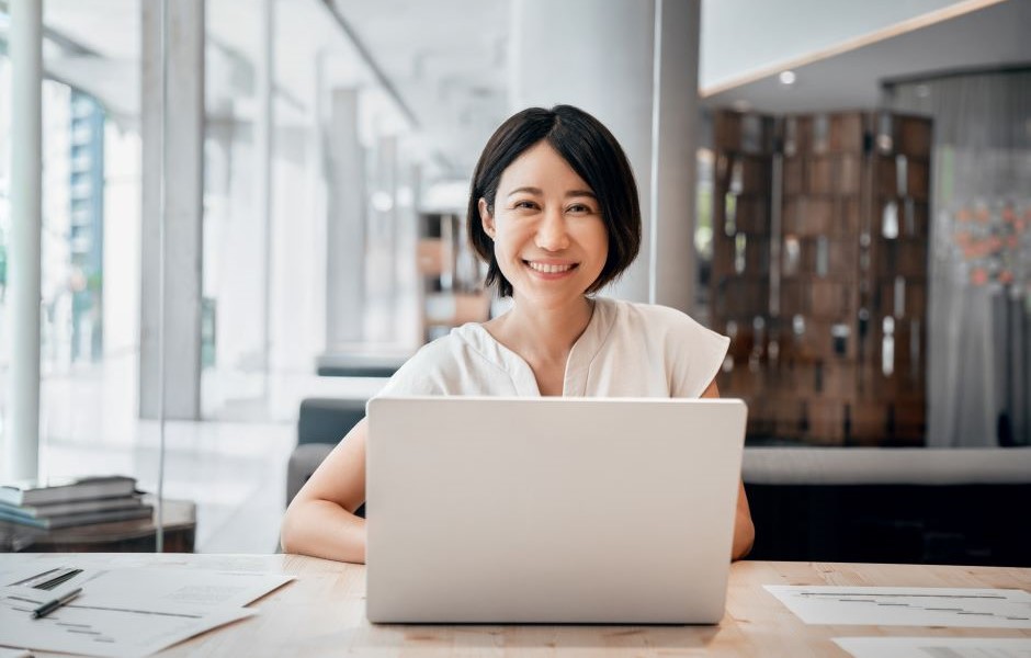 Mulher sorridente usando laptop em um ambiente de escritório moderno com iluminação natural, transmitindo produtividade e satisfação no trabalho.