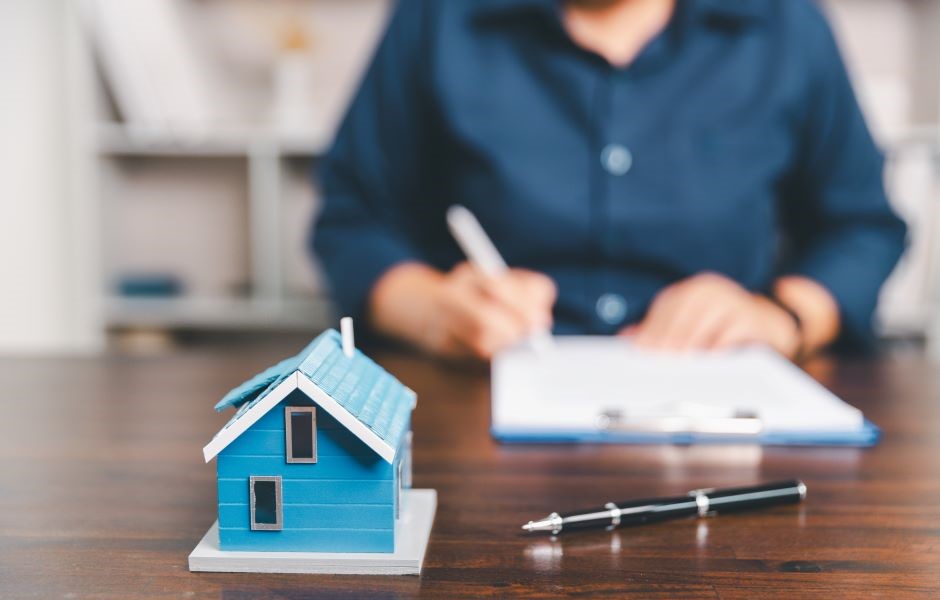 Pessoa realizando uma assinatura de contrato de aluguel de uma casa, com uma maquete de uma residência azul na mesa, simbolizando o processo de locação ou compra de imóveis.